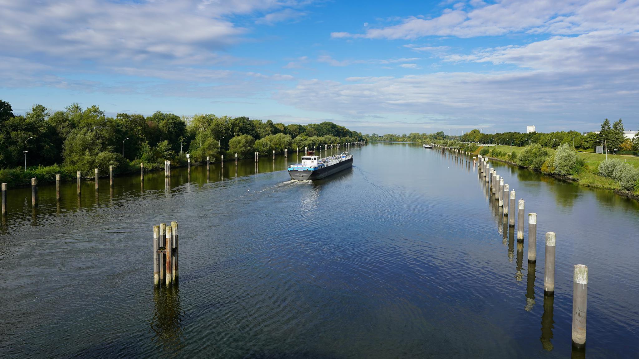 A cargo ship navigates through the serene canal in Geesthacht, Germany, under a clear blue sky.